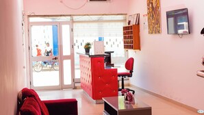 A reception area with a red sofa, a red cushioned desk, a TV mounted on the wall, and a window with blinds. - Hospedaria RaSimão 1 (Benguela)