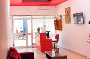 Uma área de recepção com um sofá vermelho, uma mesa com estofamento vermelho, uma televisão na parede e uma janela com persianas.