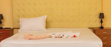 A neatly made bed with a white pillow and a folded towel, a wooden nightstand with a lamp, and a wooden dresser with drawers.