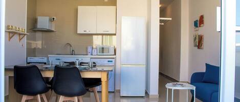 Una cocina moderna con armarios blancos, microondas y refrigerador. Hay dos sillas negras en una mesa de madera con una mesita auxiliar blanca. Se ve un sofá azul al fondo.