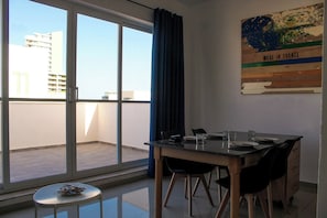 A dining area with a table and chairs, a small round table with a newspaper, and a view of buildings through large windows. - Tranquil Cityscape Penthouse (Gzira)