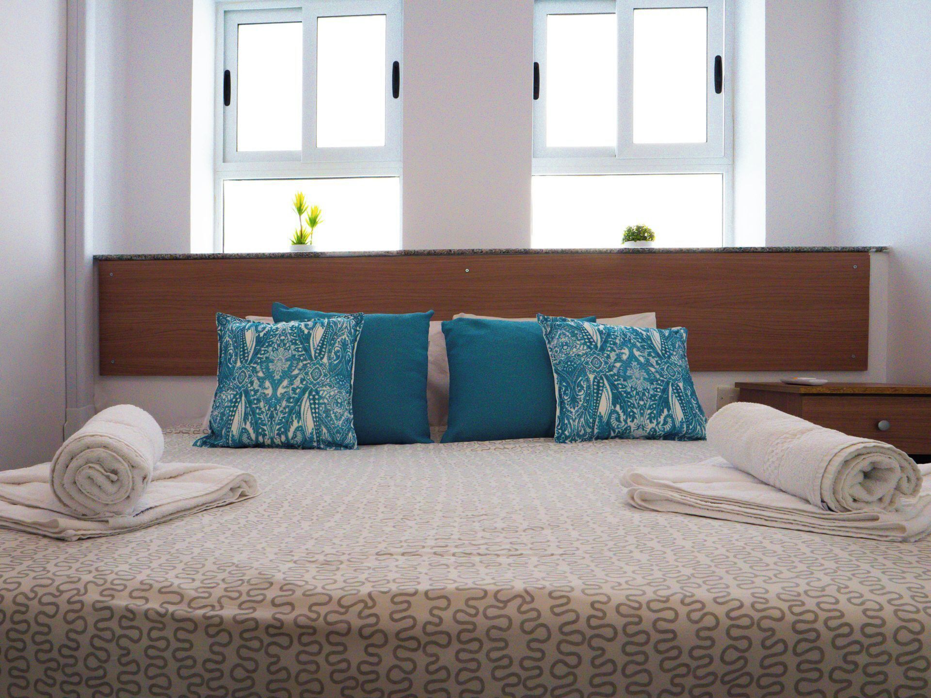 A neatly made bed with patterned bedding, two windows with blinds, and a wooden headboard.