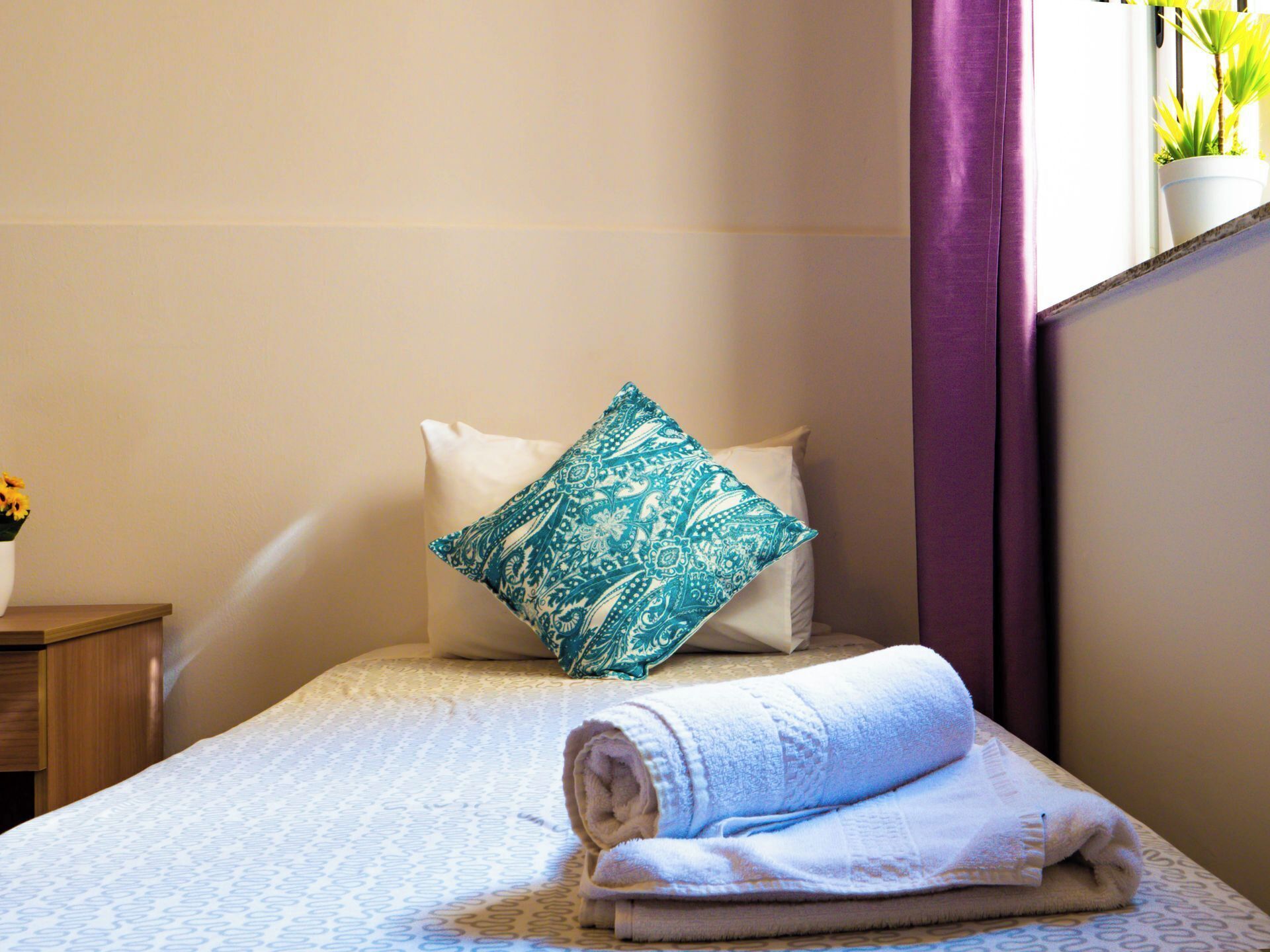 A neatly made bed with a patterned pillow, a rolled towel, and a small table with a flower vase.