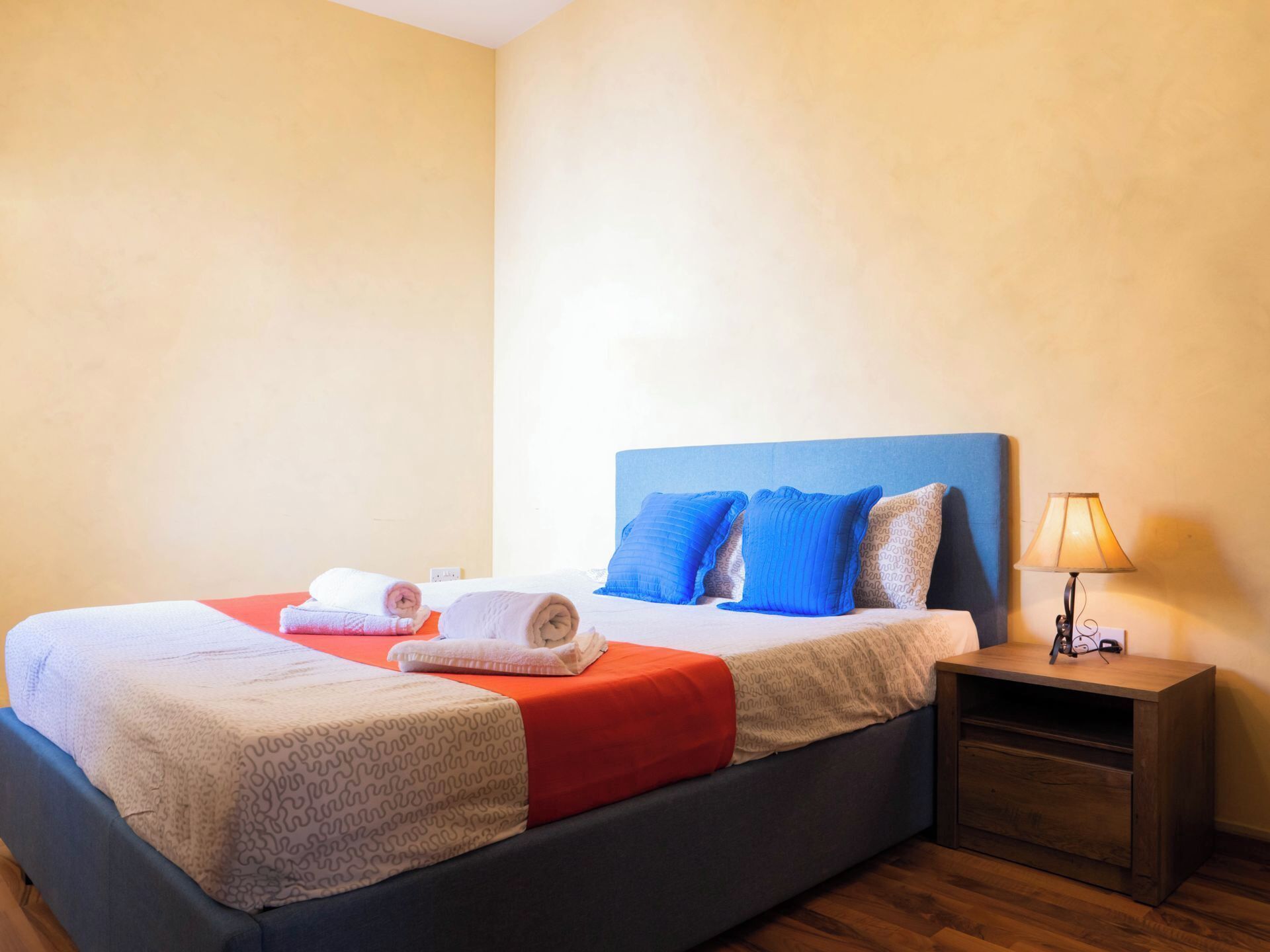 A neatly made bed with blue and white pillows, a red blanket, and two rolled towels. There is a wooden nightstand with a lamp beside the bed.