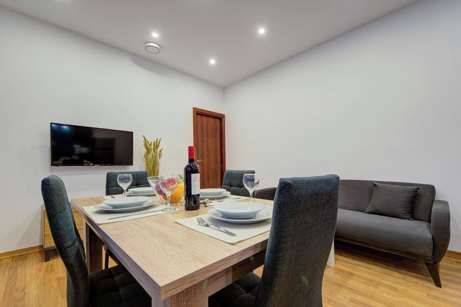 A dining area with a wooden table set for four, a bottle of wine, and a flat-screen TV on the wall.