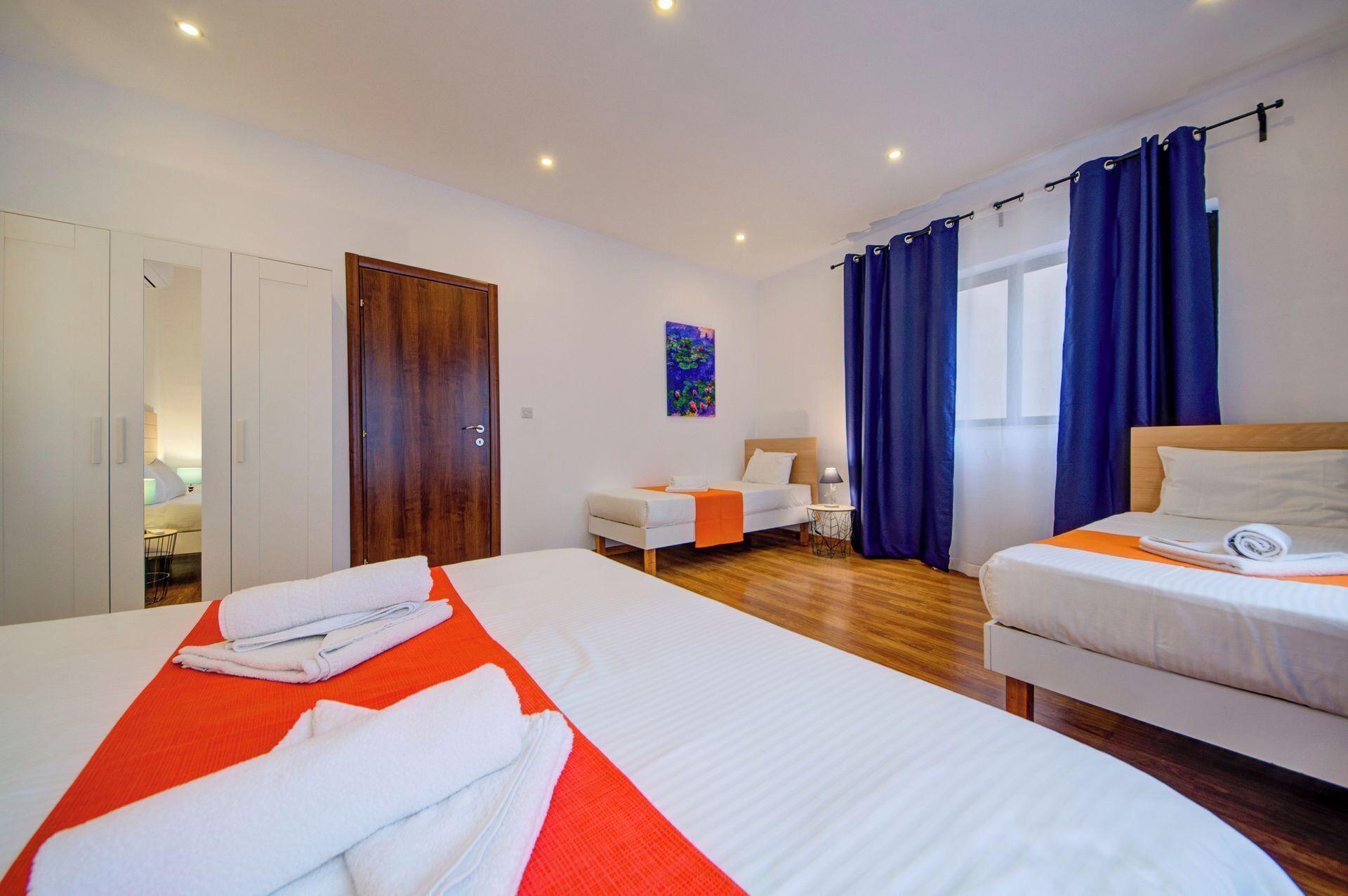 A hotel room with two beds, a wooden wardrobe, a painting on the wall, and a window with blue curtains.