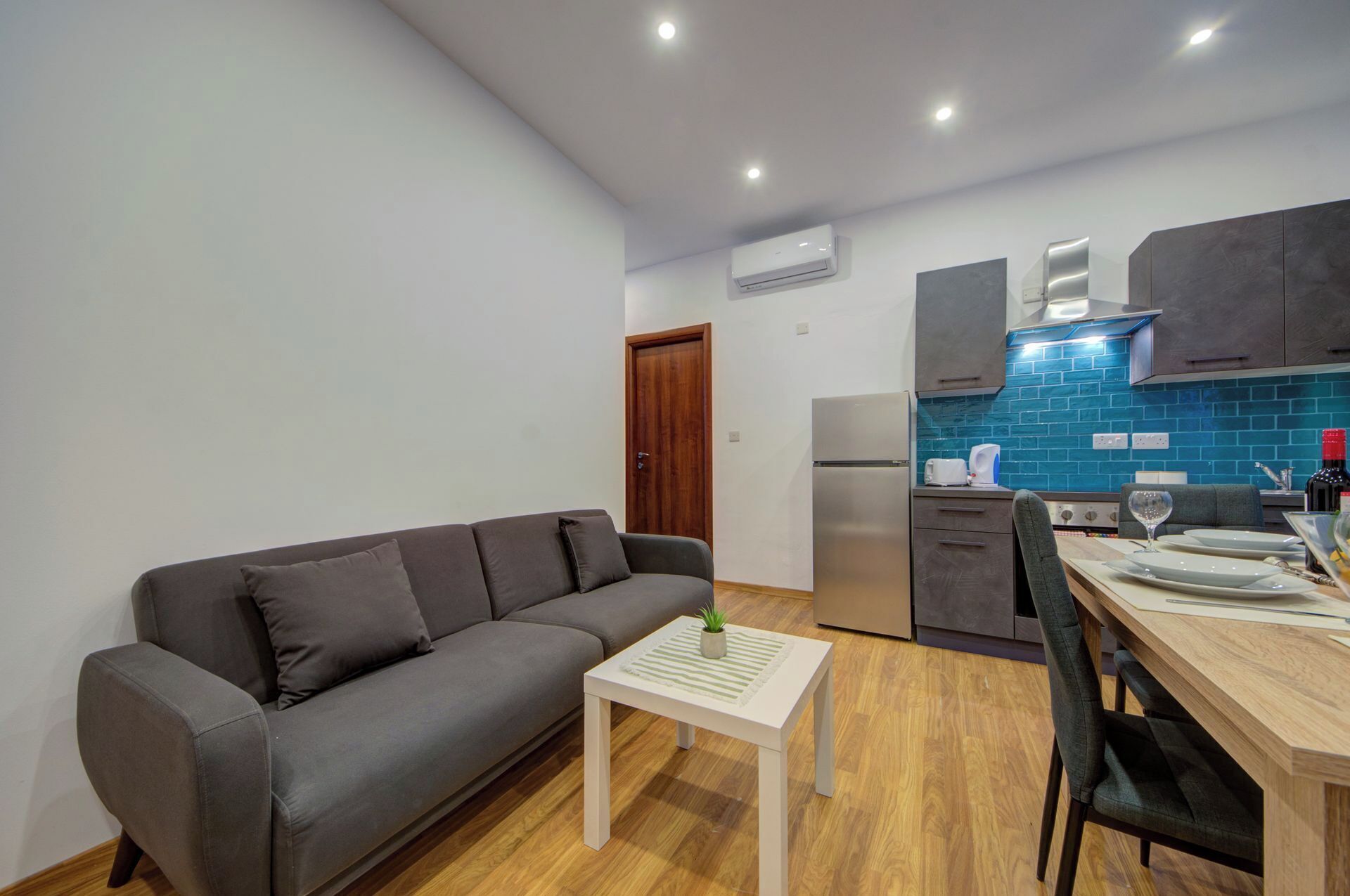 A modern living room with a grey sofa, a small white table with a plant, and a wooden dining table with chairs. The kitchen features a stainless steel refrigerator, a blue tiled backsplash, and dark wooden cabinets.