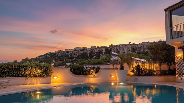 A swimming pool with a sunset view, a building with a balcony, and a hill with buildings in the background. - Villa Alexandra: Sea Views, Pool, Jacuzzi & Sauna (Mellieha)