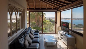 A modern living room with a black leather sofa, a white coffee table, and a TV on a white stand. The room has large windows offering a view of a body of water and greenery. - Villa Alexandra: Sea Views, Pool, Jacuzzi & Sauna (Mellieha)