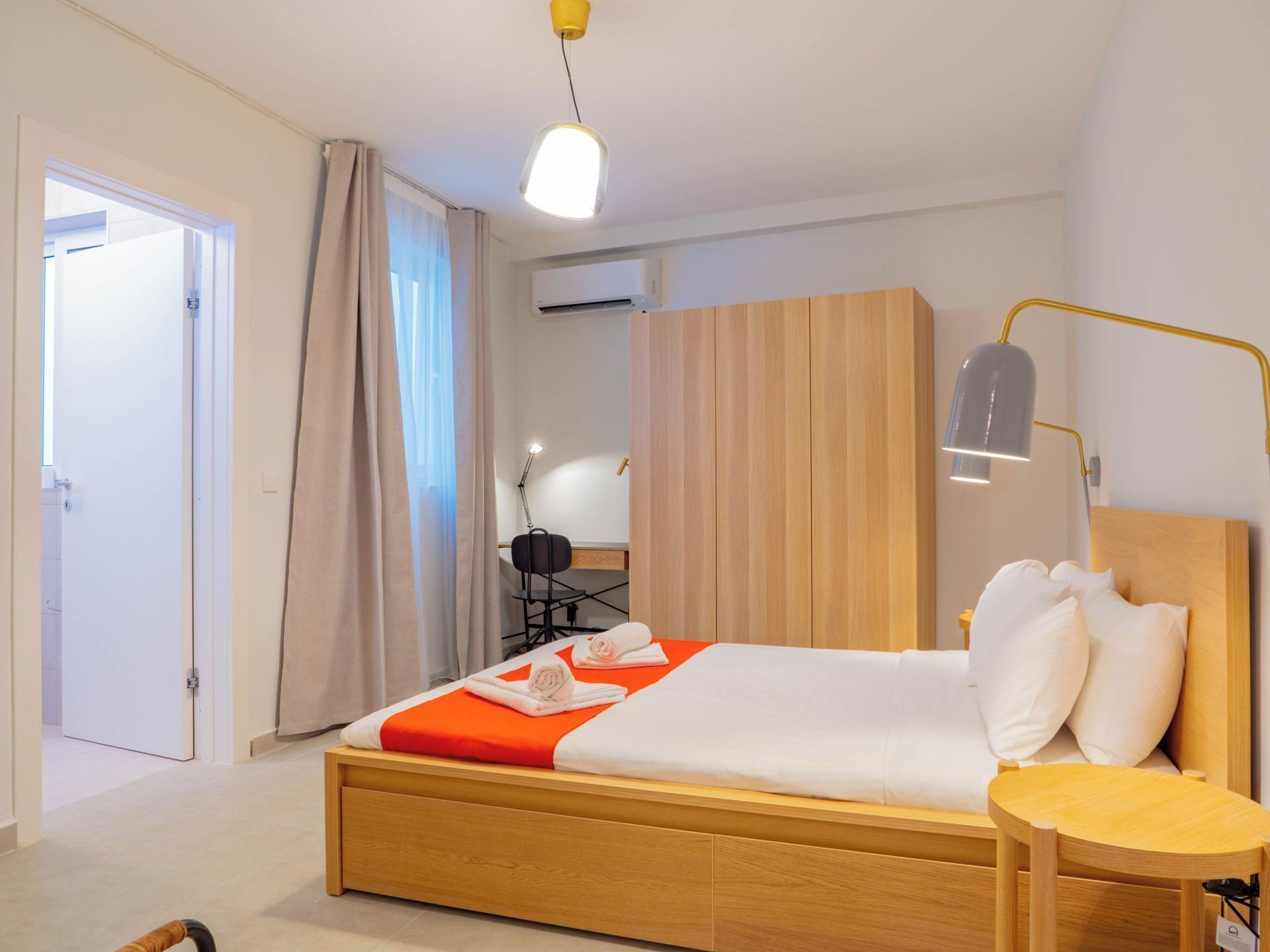 A hotel room with a wooden bed, a bedside table, a desk with a chair, an air conditioning unit, and a window with curtains.