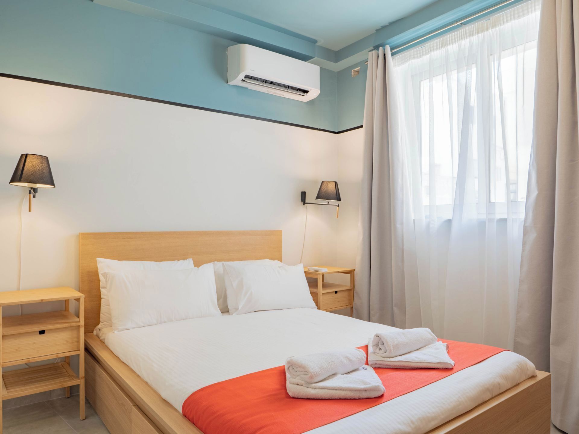 A hotel room with a wooden bed, white bedding, two bedside lamps, an air conditioning unit, and a window with sheer curtains.
