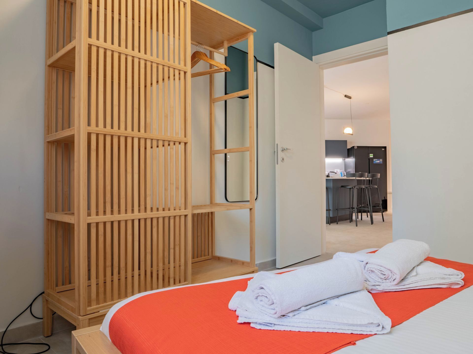 Quarto de hotel com guarda-roupa de madeira, cama com colcha laranja e toalha branca dobrada sobre a cama.