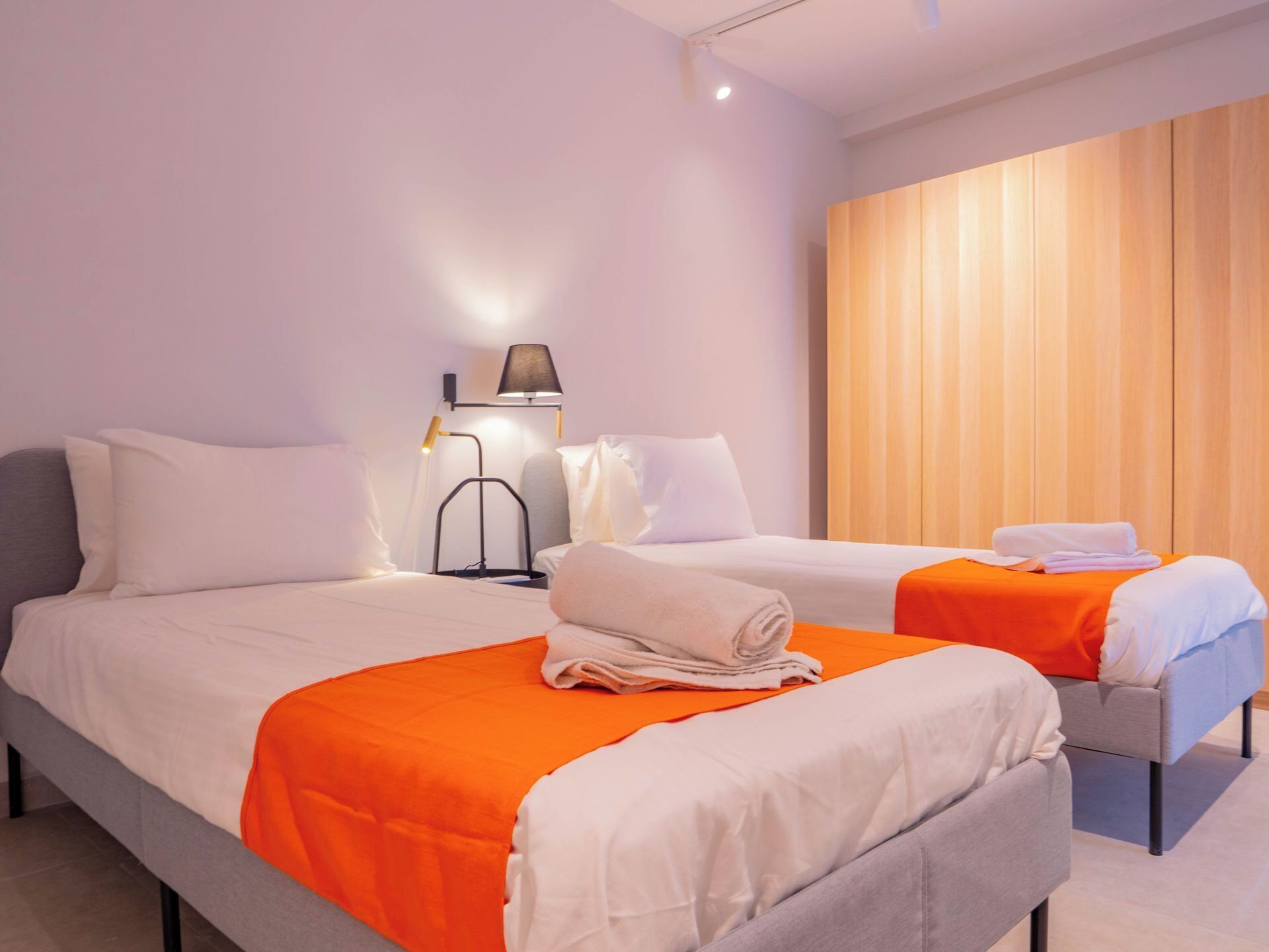 A hotel room with two beds, each with an orange bedspread and white linens. There is a nightstand with a lamp between the beds, and a wooden panel wall on the right side.