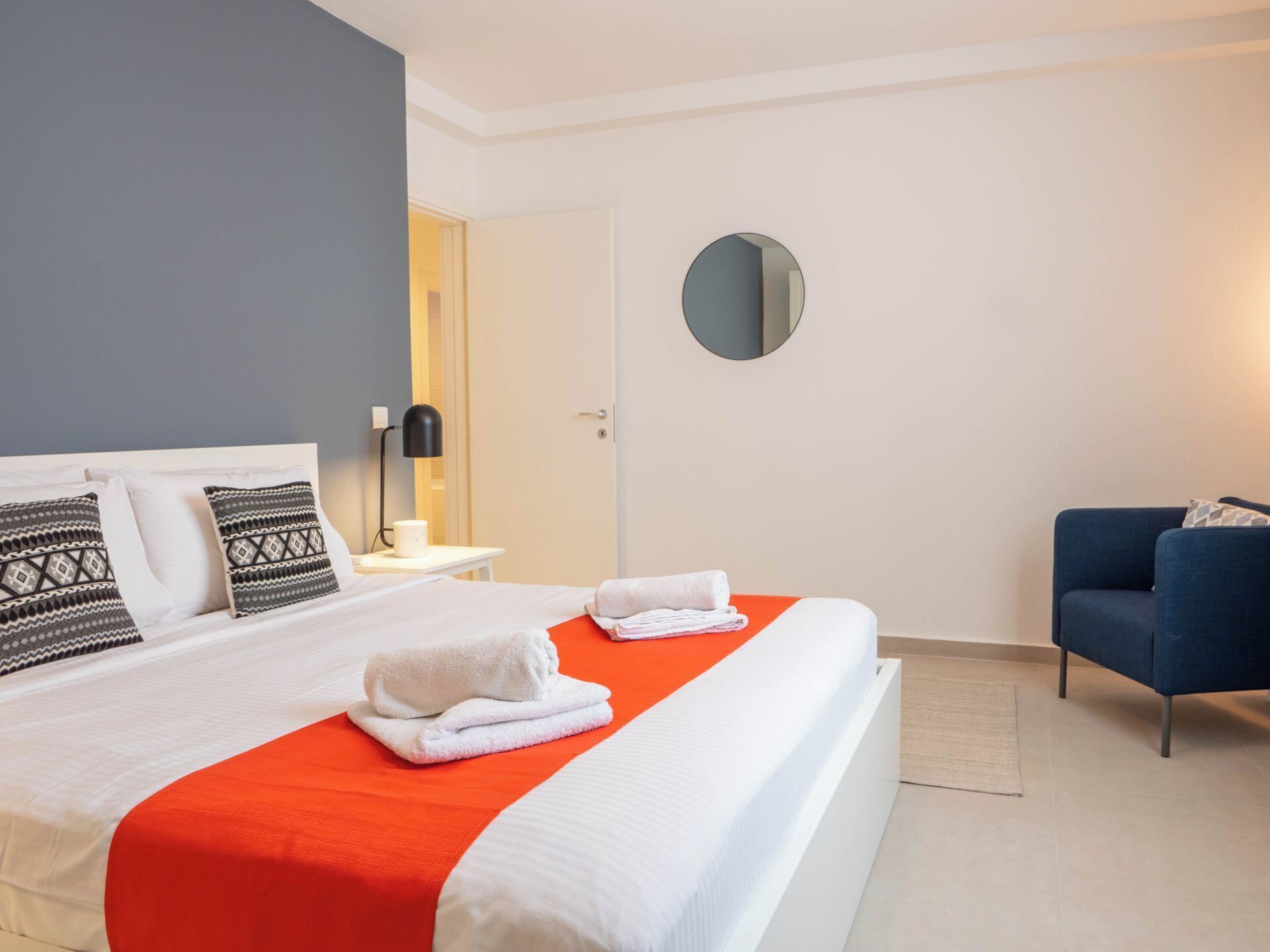 Une chambre d’hôtel avec un grand lit, une table de chevet avec une lampe, un miroir rond et un fauteuil bleu.