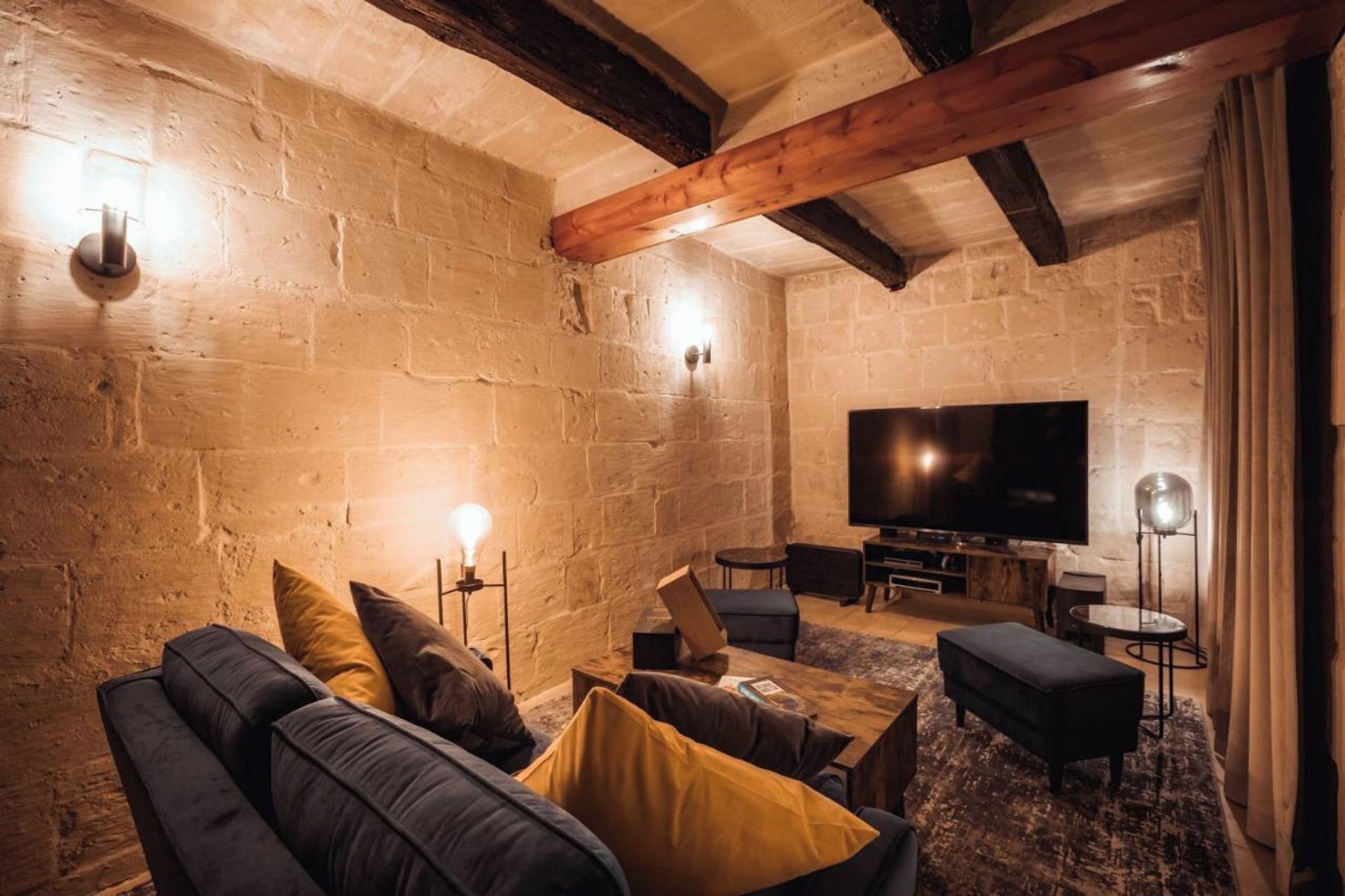 A cozy living room with a stone wall, wooden beams, a sofa, a coffee table, and a flat-screen TV.