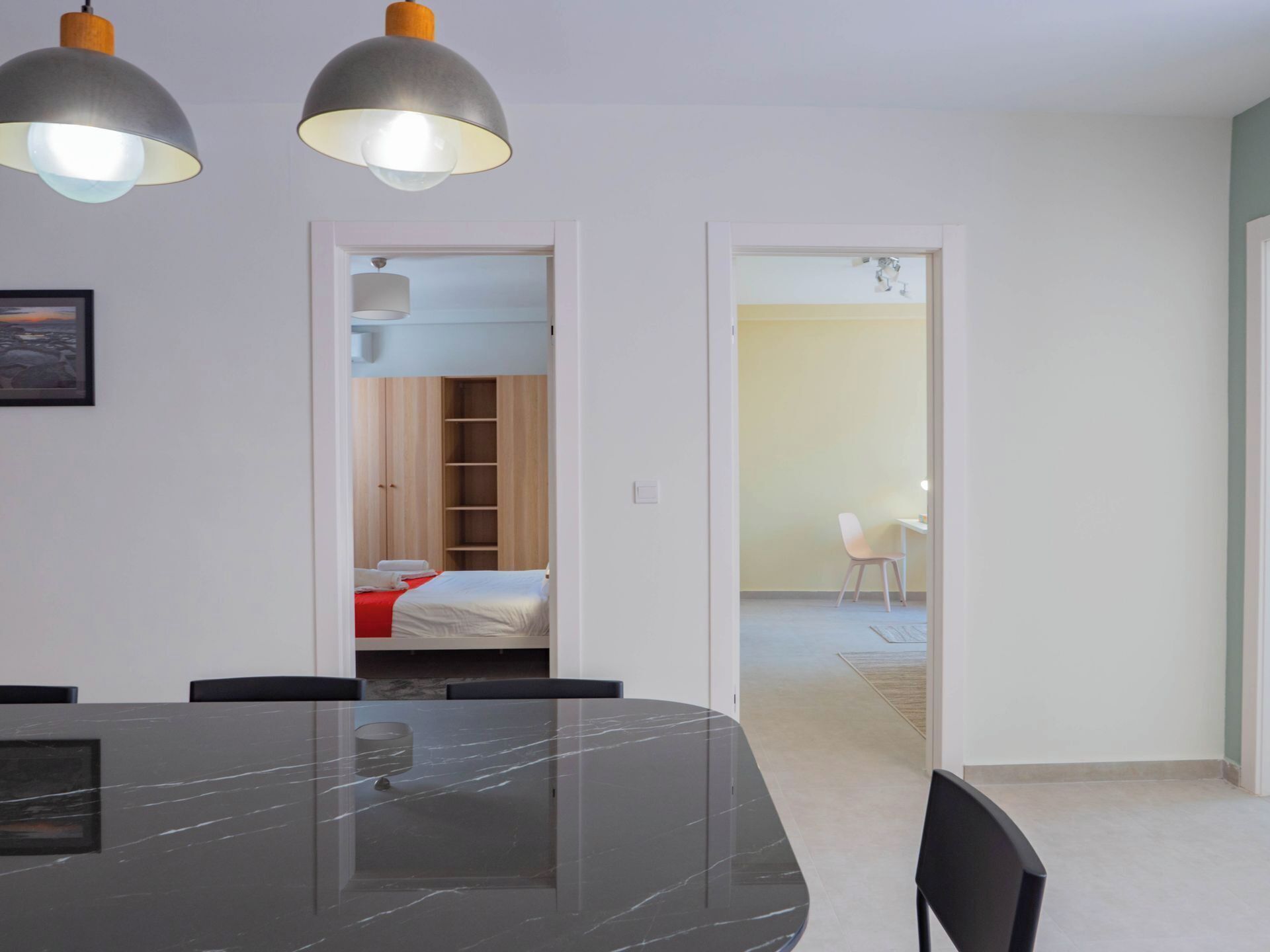 Une chambre moderne avec une table en marbre, deux lampes suspendues, un lit recouvert d’une couverture rouge, une armoire en bois et une chaise dans la pièce adjacente.