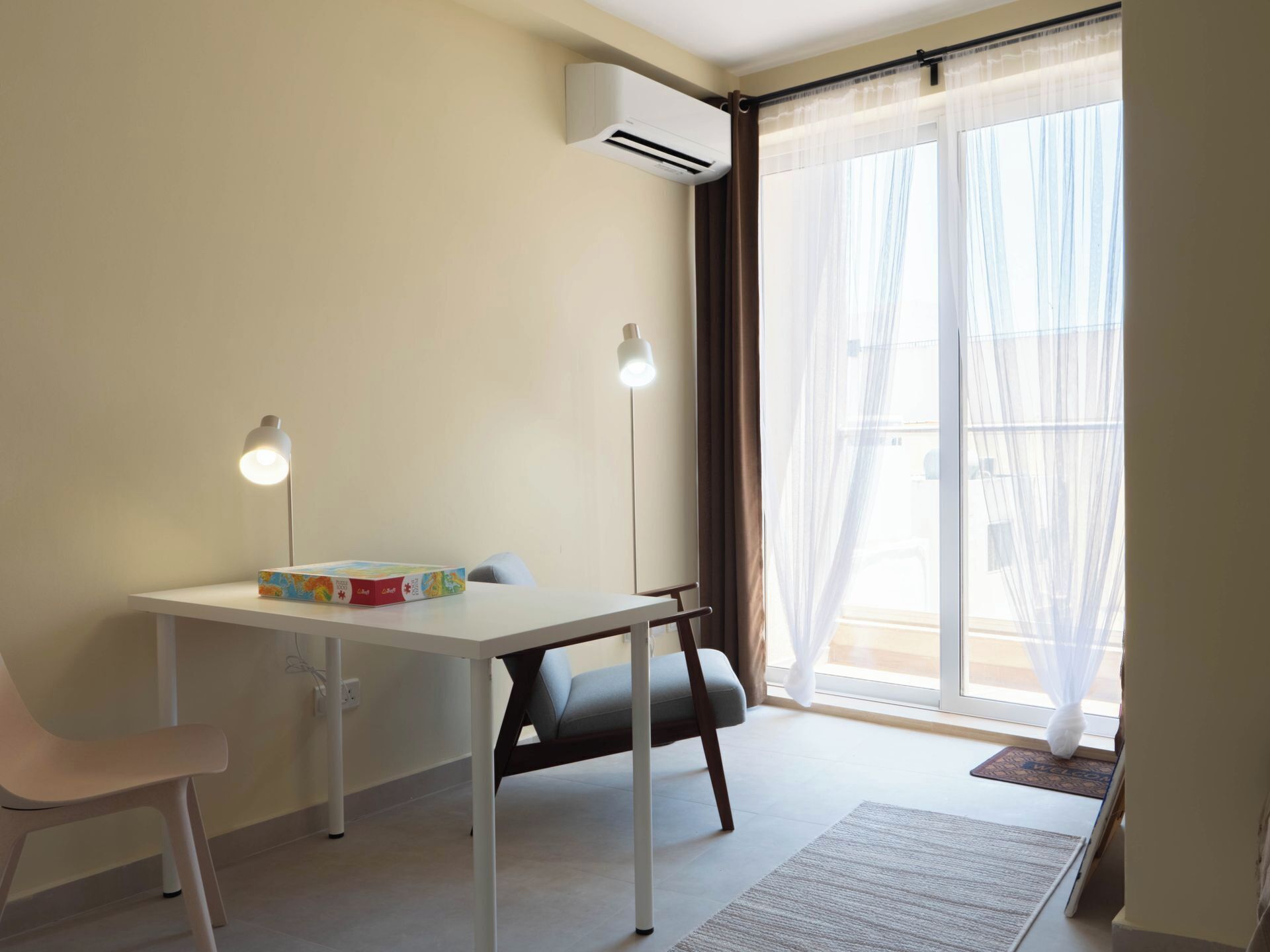 Una habitación con una mesa blanca, dos lámparas, una silla, un ventanal con cortinas translúcidas y un equipo de aire acondicionado.