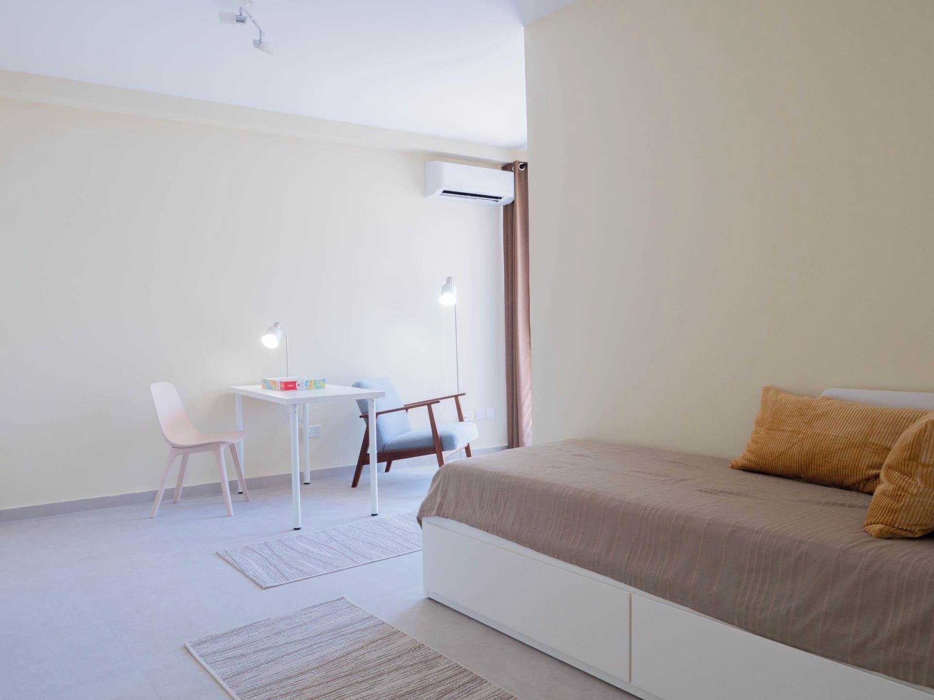 A bedroom with a bed, a desk, a chair, and an air conditioning unit.
