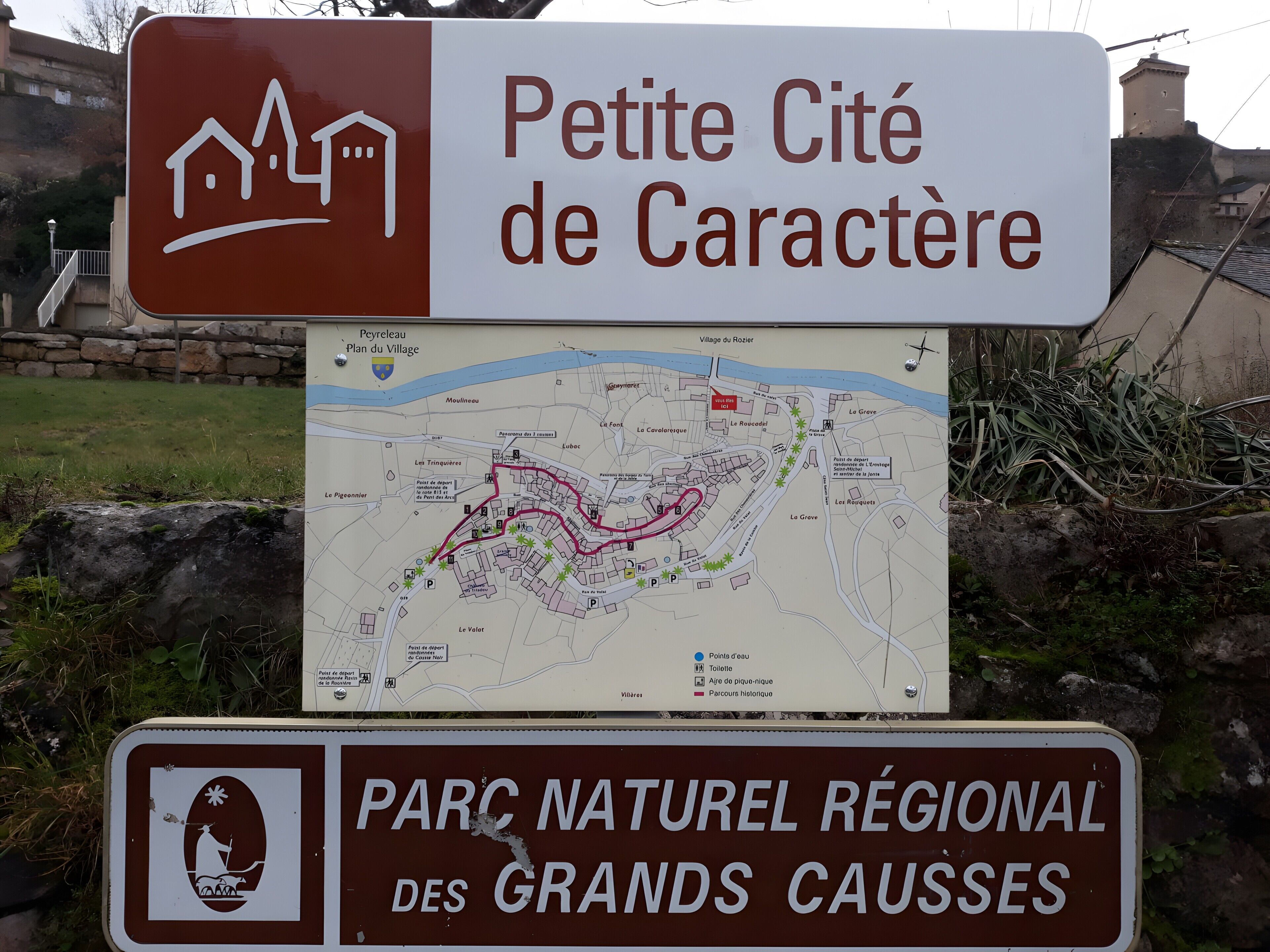 Un cartel con un mapa y texto que indica 'Petite Cité de Caractér' y 'Parc Naturel Régional des Grands Causses'.