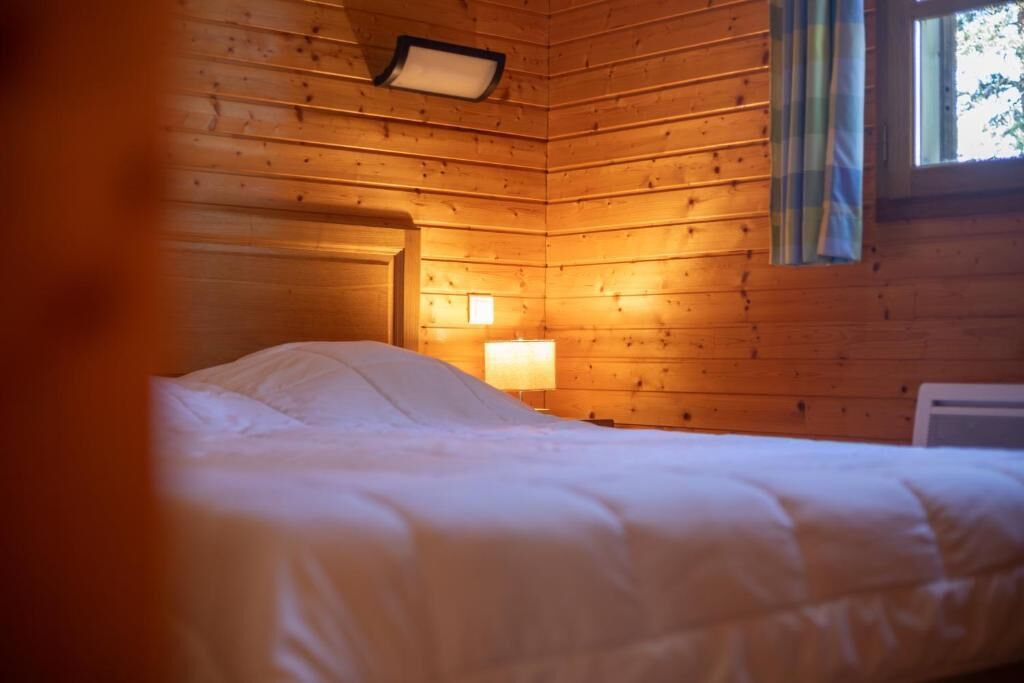 A wooden cabin room with a bed, a nightstand with a lamp, and a window with curtains.