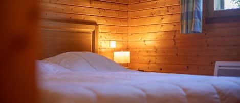 A wooden cabin room with a bed, a nightstand with a lamp, and a window with curtains.