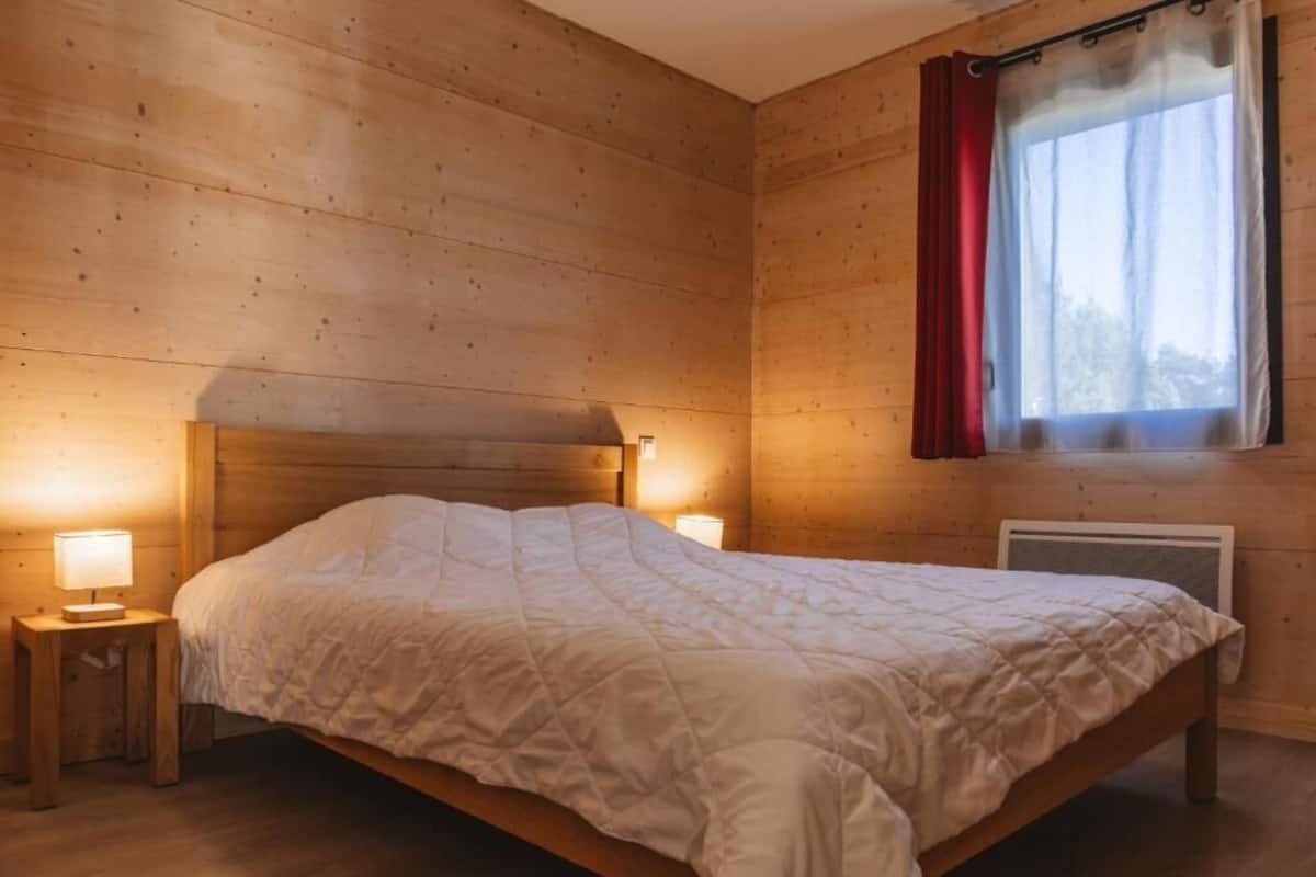 A wooden-paneled bedroom with a bed, two bedside lamps, and a window with a red curtain.