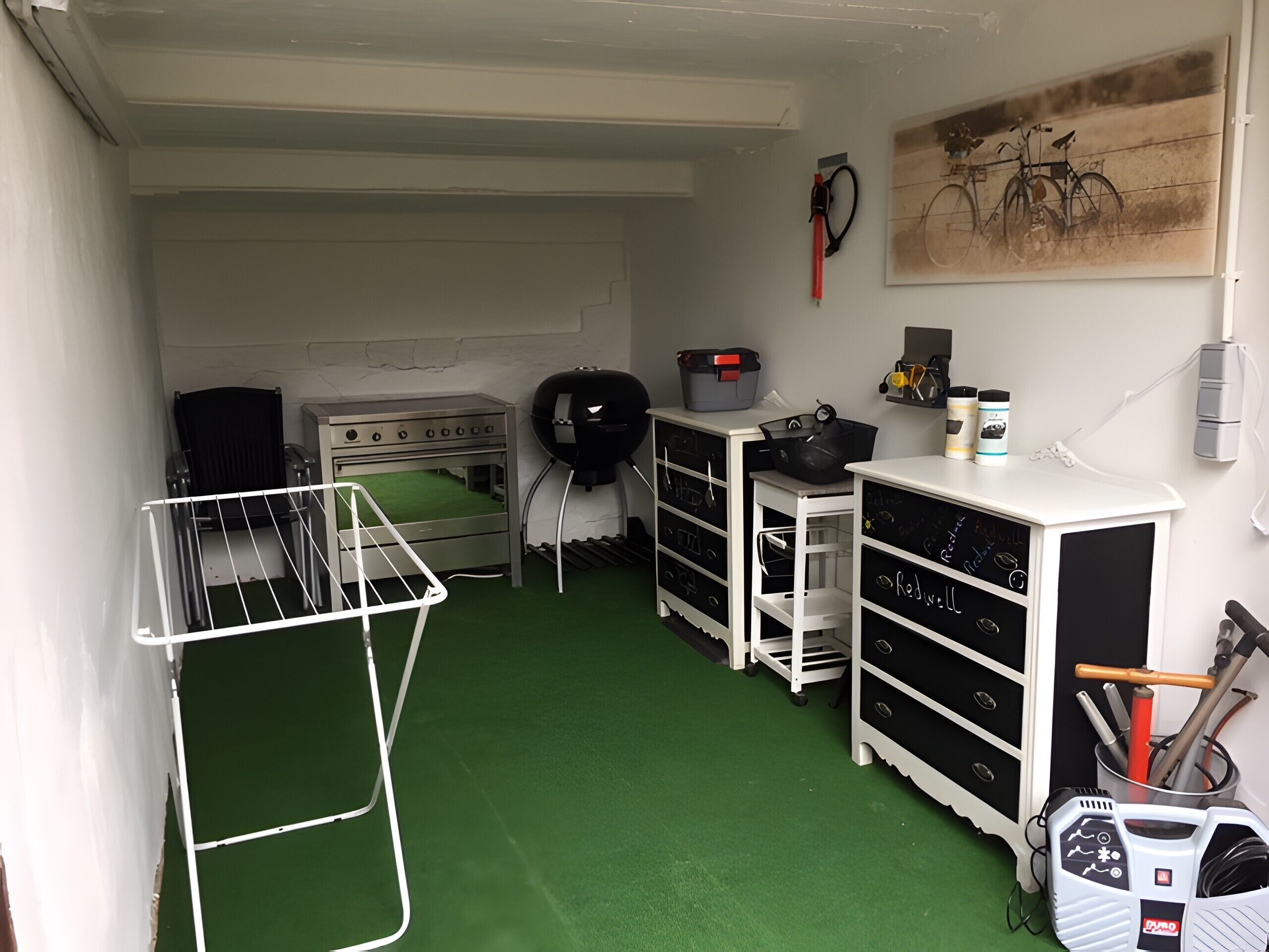 A compact kitchen with a gas stove, drying rack, storage cabinets, and a bicycle painting on the wall.