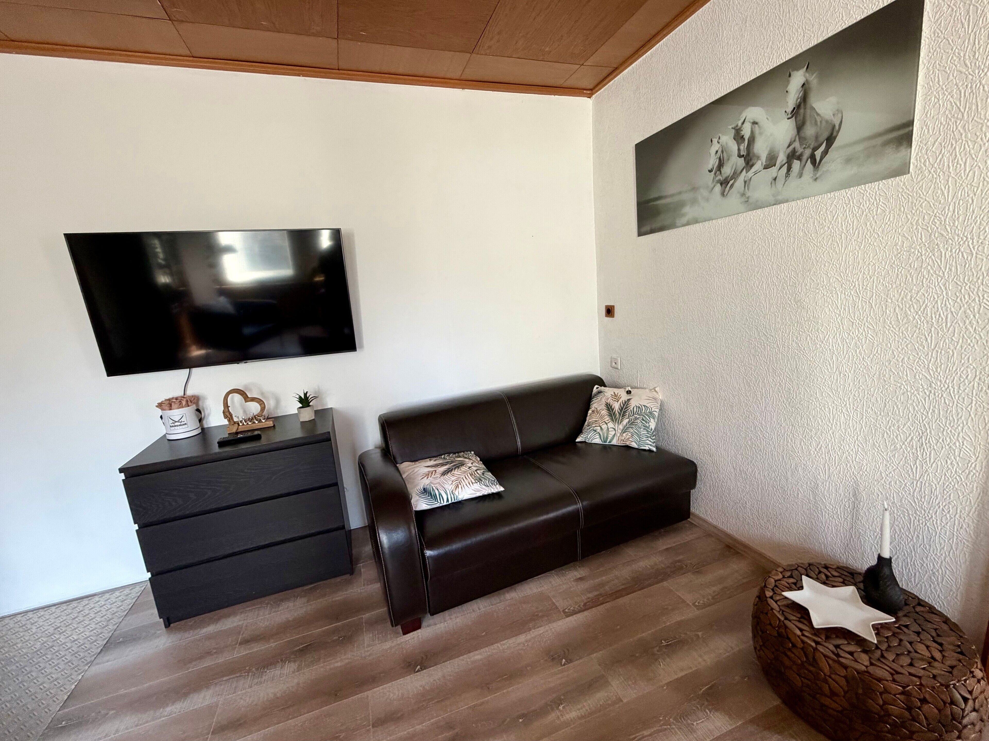 A living room with a flat-screen TV, a dark leather sofa, a small side table, and a wall-mounted painting.