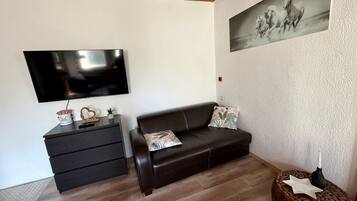 A living room with a flat-screen TV, a dark leather sofa, a small side table, and a wall-mounted painting.