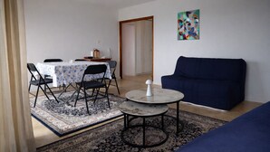 Uma sala de estar com um sofá azul, uma mesa de centro redonda, uma área de jantar com mesa e cadeiras, e um quadro abstrato colorido na parede.