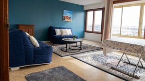 Uma sala de estar com um sofá azul, uma mesa de centro redonda, uma mesa pequena com toalha de mesa florida e uma vista através de grandes janelas.