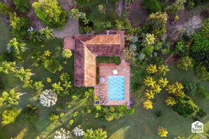 An aerial view of a house with a swimming pool, surrounded by lush greenery and palm trees.