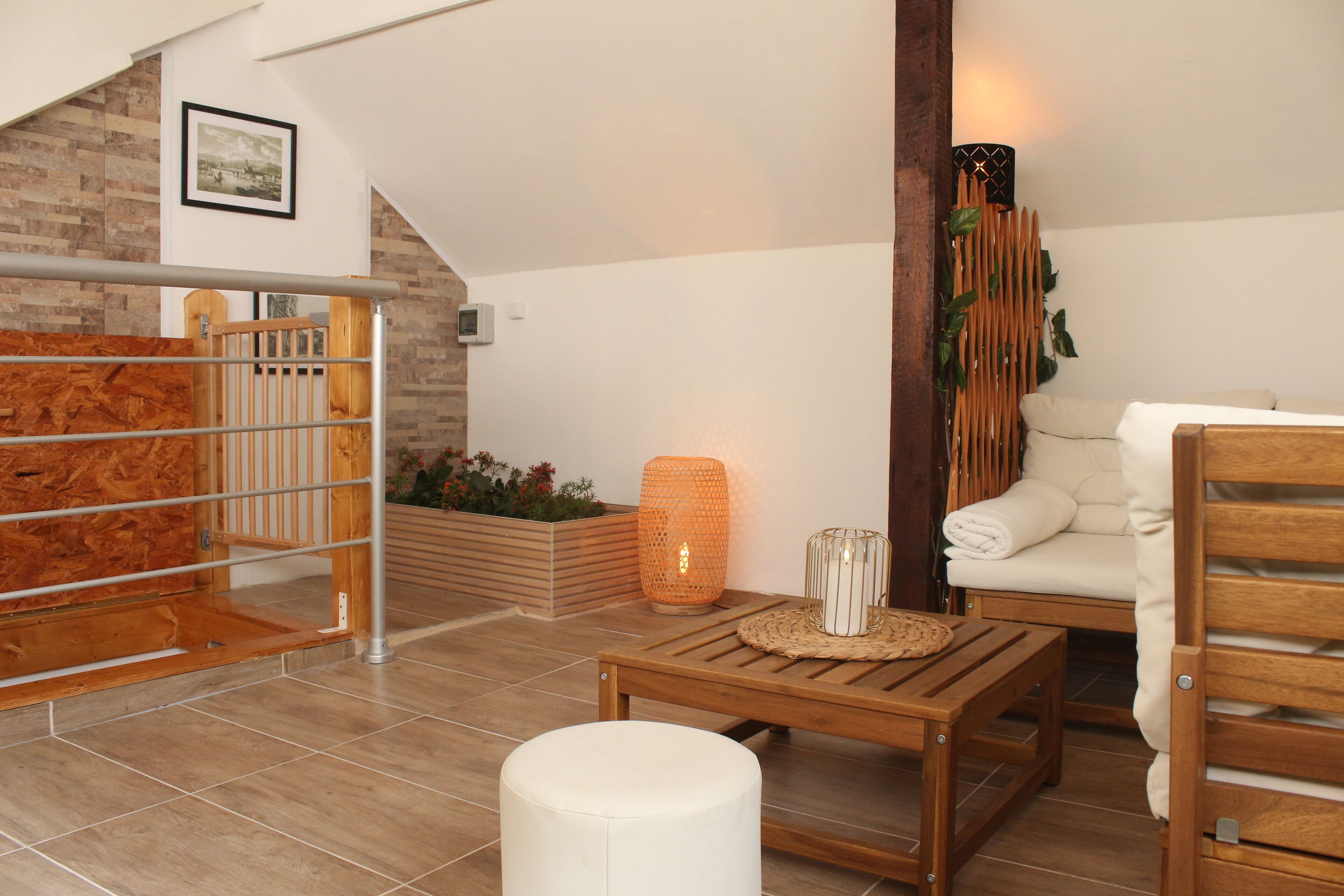 Un salón moderno con un banco de madera, un reposapiés blanco y una mesa de madera. Se ve una escalera con barandilla de madera que conduce a un nivel superior. Hay una planta y un adorno en la pared.