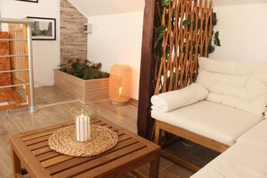 Ein Holztisch mit gewebter Tischunterlage und Kerze, ein weiß gepolstertes Sofa, ein Holzpflanzgefäß mit Blumen und ein gewebter Laterne.