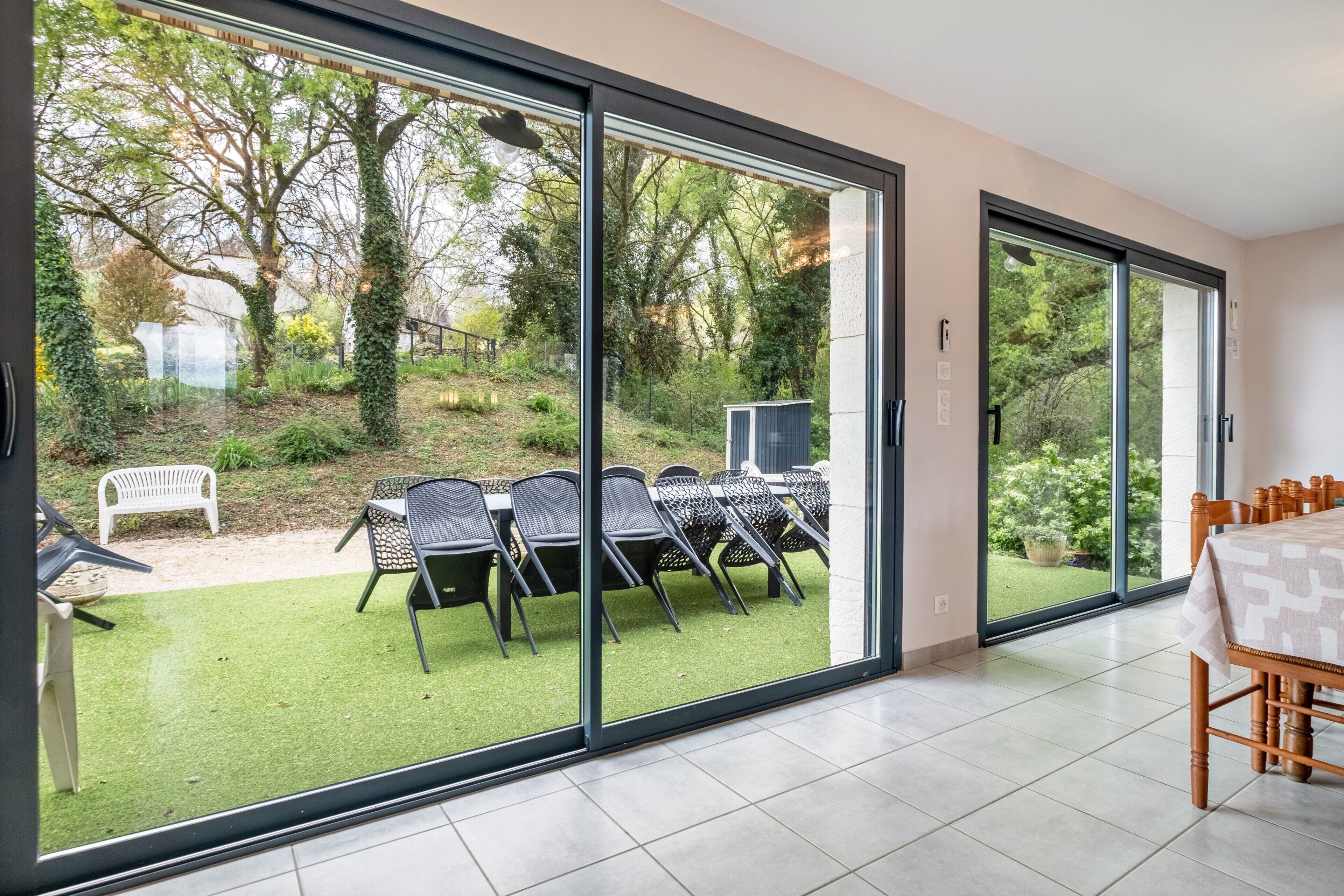A room with a large sliding glass door leading to an outdoor area with a table and chairs, a bench, and a garden view.