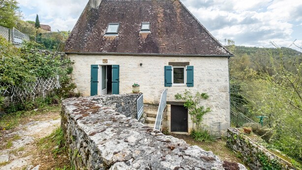 A stone wall leading to a white building with a steep roof and green shutters.
