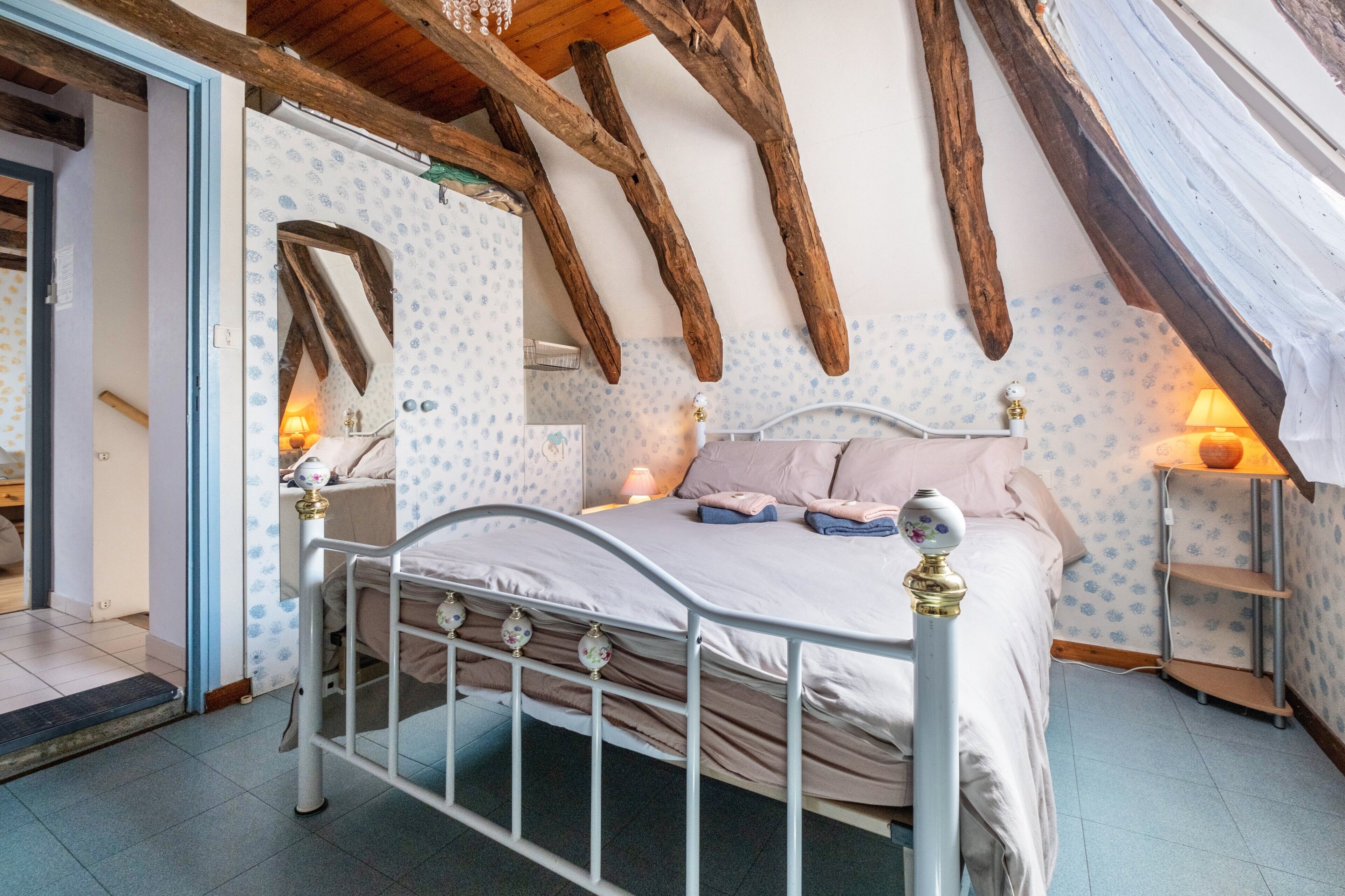 A bedroom with a bed, wooden beams, patterned wallpaper, and a nightstand with a lamp.