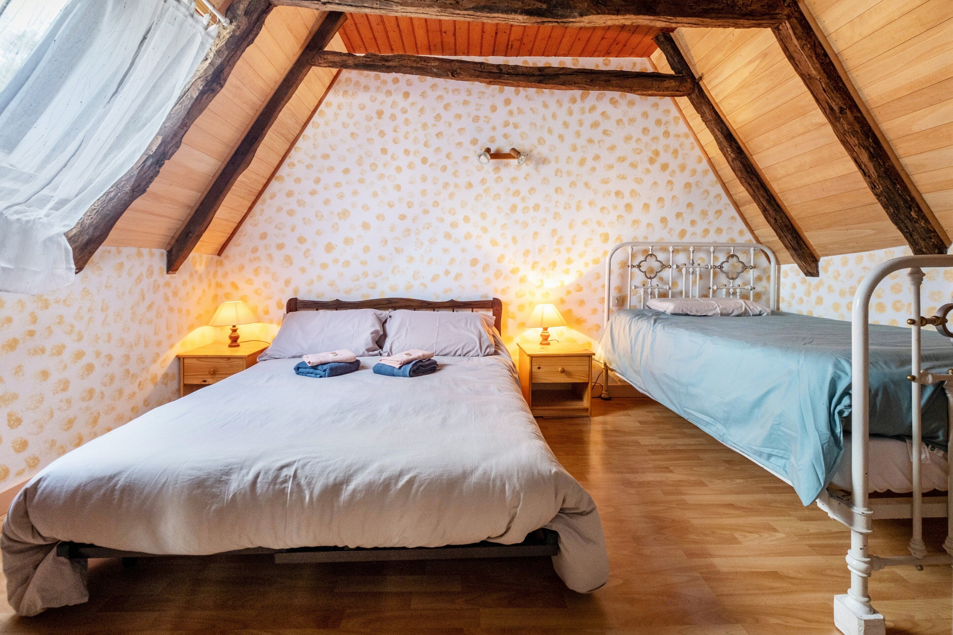A bedroom with a large bed, a smaller bed, wooden beams, patterned wallpaper, and two bedside lamps.