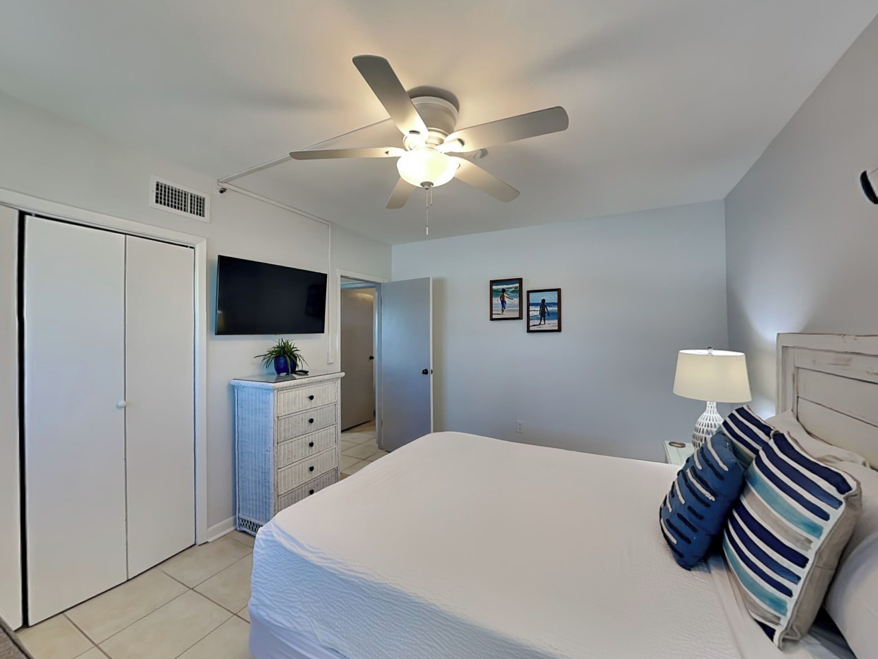 Une chambre à coucher avec un lit, un ventilateur de plafond, un téléviseur fixé au mur et une porte donnant sur une autre pièce.