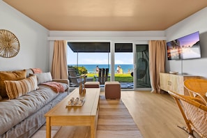 A modern living room with a sofa, coffee table, and wicker chair, a flat-screen TV, and a view of the ocean. - The Golden Hour - Ocean Front (San Diego)