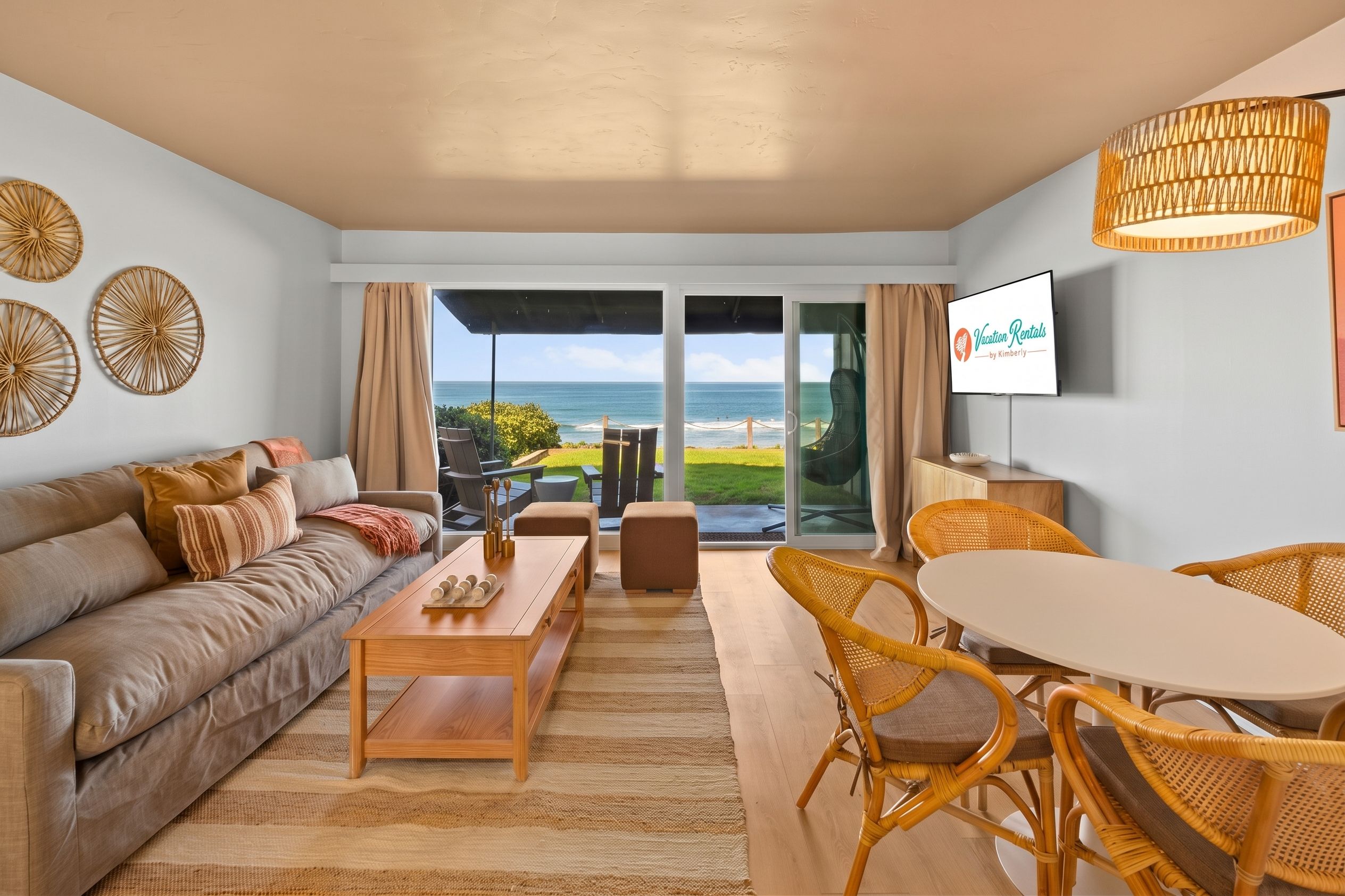 A living room with a sofa, coffee table, and wicker chairs, a television, and a view of the ocean.