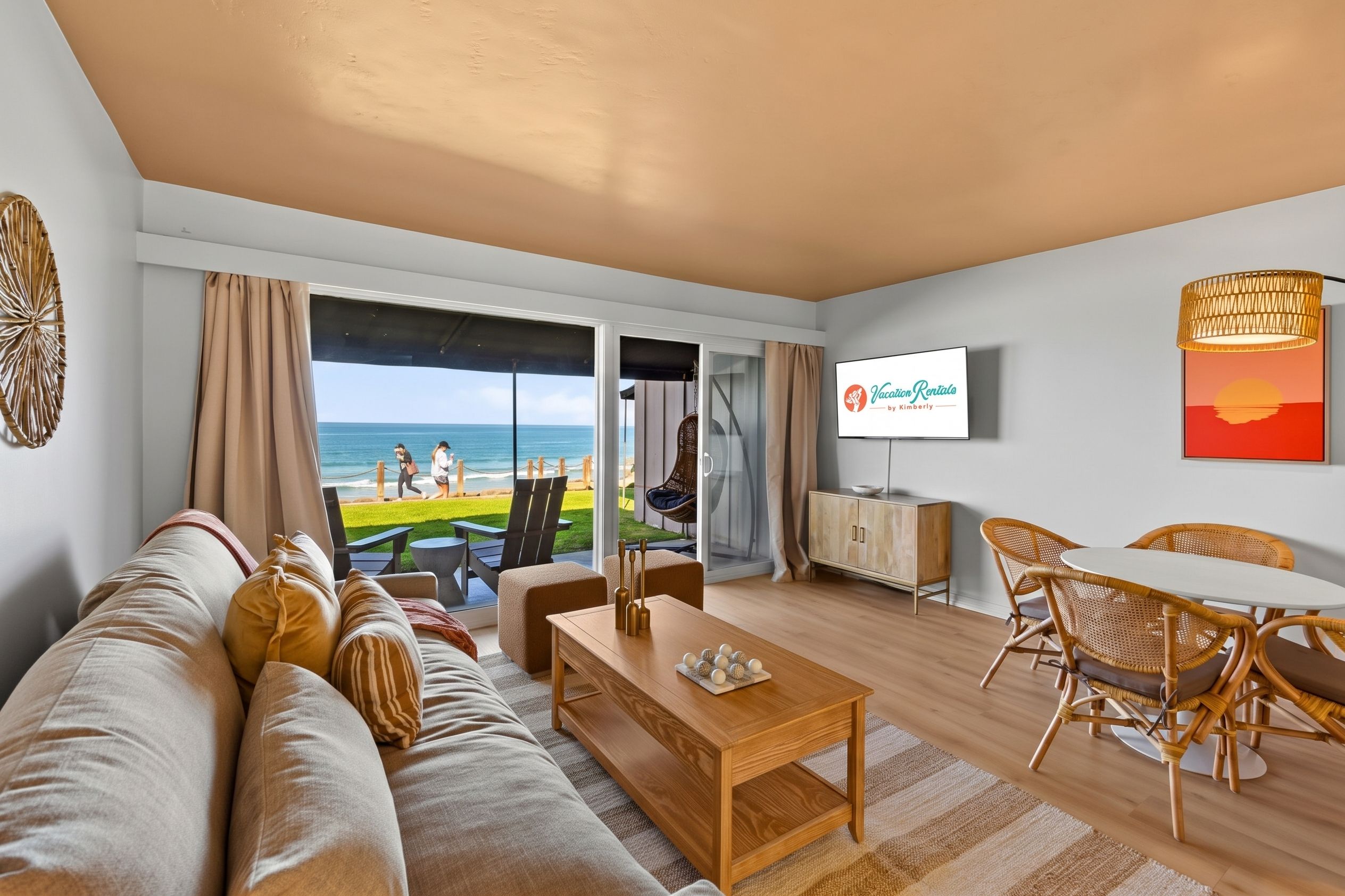 A modern living room with a large window overlooking the beach, a flat-screen TV mounted on the wall, and a dining area with wicker furniture.