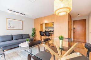 Un salon moderne avec un canapé, une table à manger en verre et des chaises. On aperçoit à l’arrière-plan un coin cuisine avec des meubles en bois et une porte.