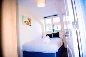 Ein Hotelzimmer mit Bett, einem Fenster mit Blick auf Gebäude, einem Bild an der Wand und einer Tür rechts.