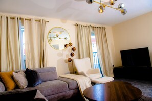 Ein Wohnzimmer mit einer Couch, einem Sessel und einem runden Couchtisch. Rechts befindet sich ein Fernseher und an der Wand ein Spiegel mit dekorativer Beleuchtung.