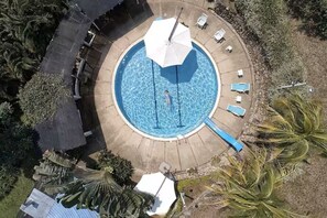 Luftaufnahme eines kreisförmigen Schwimmbeckens mit einer Person im Wasser, umgeben von Liegestühlen und Sonnenschirmen.