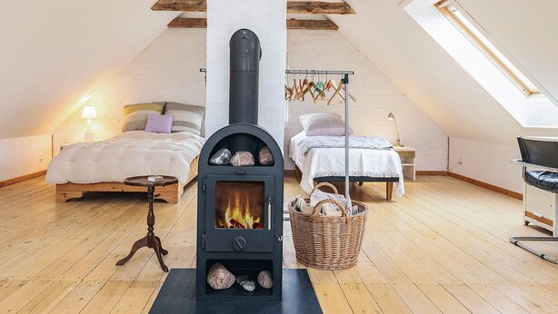 A cozy attic bedroom with a central wood-burning stove, two beds, a small table, and a hanging clothes rack. - 6 Person Holiday Home in Stege (Stege)