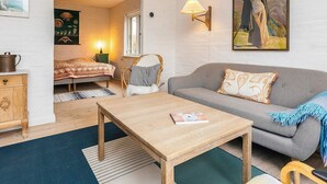 A room with a bed, a grey sofa, a wooden table, a wicker chair, a lamp, and a painting on the wall. - 6 Person Holiday Home in Stege (Stege)