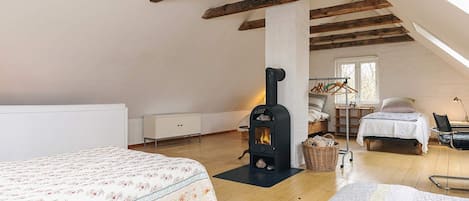 A cozy attic bedroom with a wooden ceiling, a bed with a floral bedspread, a black wood-burning stove, and a window providing natural light.