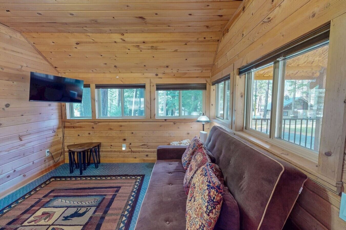 A cabin interior with a wooden ceiling, a sofa, a TV, and large windows.